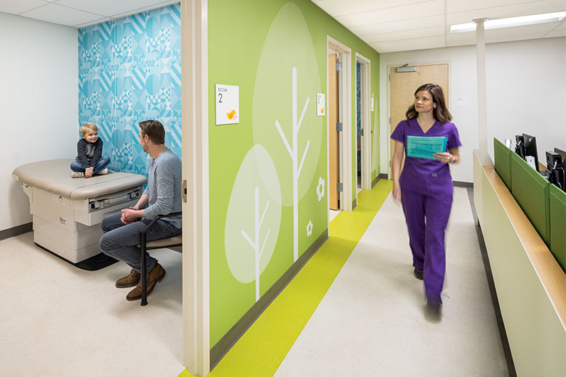 Healthcare Design - Nurse walking with blue file into room with father and son waiting for appointment