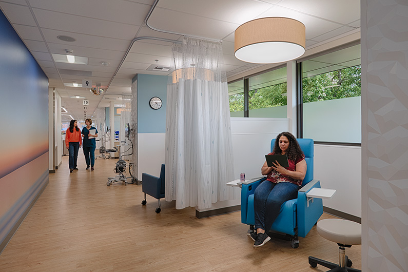 Healthcare design for critical care - Woman seated in oncology infusiton bay reading a book with two staff members walking down the hallway near her
