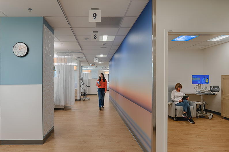 Healthcare design for critical care - Interior photo of woman sitting in private infusion room and a staff member walking down the hallway outside the room. The hallway has a nature-inspired mural on the wall
