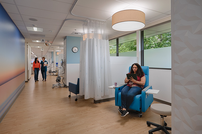 Woman seated in oncology infusiton bay reading a book with two staff members walking down the hallway near her
