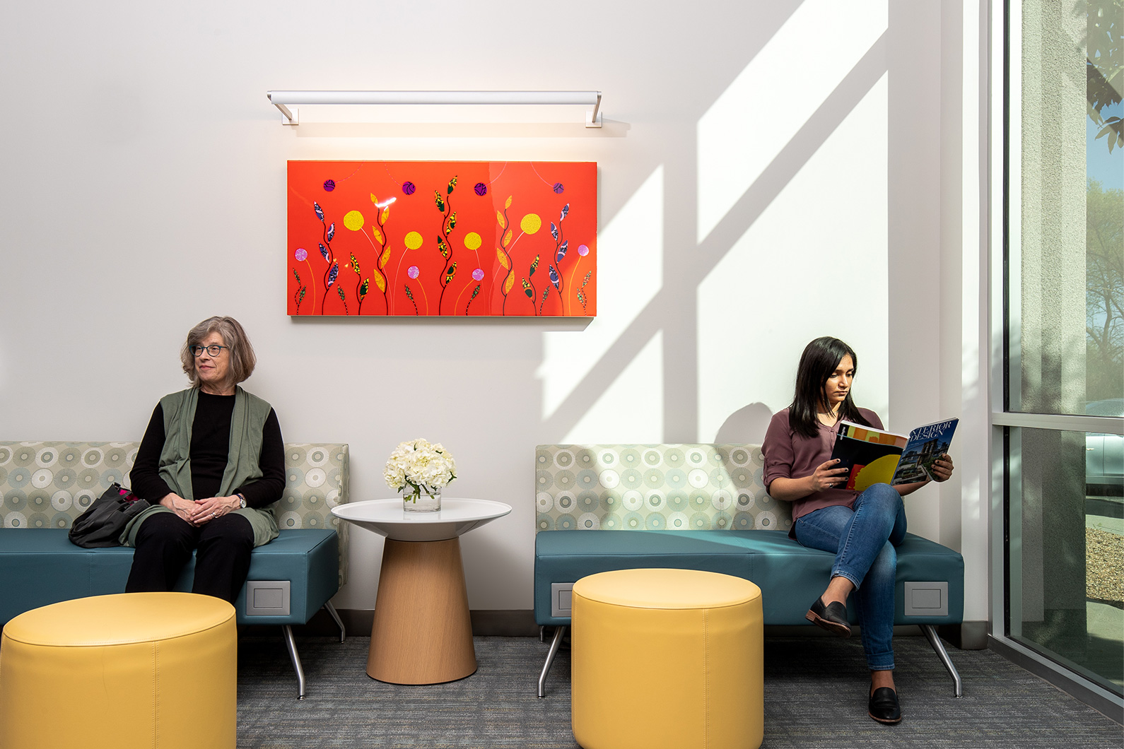 Two women seated in waiting alcove with soft light coming in from window. Colorful wall art and residential furniture complete the space